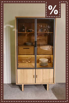 Glasvitrine aus hellem Holz auf Füßen stehend und Türen mit schwarzem Rahmen vor einer hellen Wand