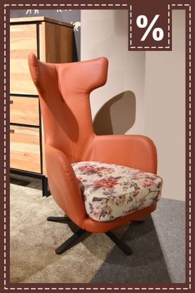 Orangener Ledersessel mit floral gemusterter Sitzfläche und schwarzem Sternfuß. Prozentzeichen.