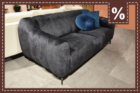 Dunkelgraues Sofa mit sanft geschwungenen Armlehnen und zwei blauen Kissen. Prozentzeichen.