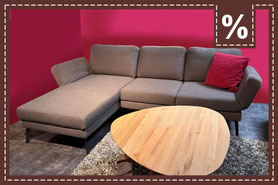 Graues L-Sofa mit ovalem Couchtisch aus Holz auf einem Teppich vor einer roten Wand. Prozentzeichen.