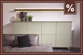 Niedriges Sideboard in hellem Grün mit Deko, darüber beleuchtete Wandleiste und ein graues Sofa. Prozentzeichen.