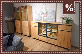 Sideboard aus hellem Holz mit Glastüren und Beleuchtung, dazu zwei hohe Vitrinen. Prozentzeichen.
