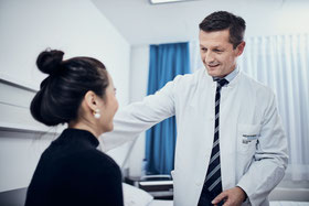 Ein Arzt in einem weißen Kittel mit Namensschild und Krawatte lächelt und spricht mit einer auf einem Bett sitzenden Patientin in einer Klinik.