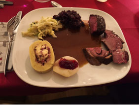 weisser Teller, darauf aufgeschnittene hellrosa gebratene Stücken von Hirschrückfilet und Rehkeule auf feiner dunkler Wildsauce neben Rahmwirsing, Rotkohl, einem Wickelkloss mit Schinkenspeck und einer Birne mit Preiselbeeren