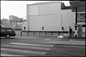 Photographie noir et blanc. Un couple gonfle une grande piscine gonflable dans une pompe à essence d'un magasin Colruyt. On voit la voiture avec le coffre ouvert sur la gauche de l'image. Bruxelles