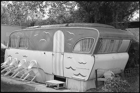 Photographie noir et blanc. Belle caravane aux angles arrondis et décorée de mouettes et de vagues peintes en un trait sur le haute et le bas. 4 chaises en plastique sont posées en biais le long de la caravane. Camping Cosmos à Westende en Belgique. 