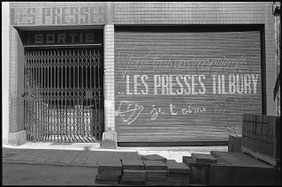 Photographie noir et blanc. Les presses Tilbury, façade avec volet peint, sur lequel on voit un tag "je t'aime" avec un coeur et une flèche. A gauche, le garage avec une grille métallique fermée. Rue Sans Souci, Ixelles, Bruxelles.