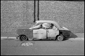 Photographie noir et blanc. Une vieille voiture, cabossée, avec 2 pneus crevés, abandonnée dans une rue et garée le long d'un mur en brique. Une bache en plastique sort par les deux fenêtres latérales coté gauche.