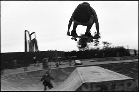 Photographie noir et blanc. Vue de dos d'un skateur inconnu effectuant un double grab en transfer dans le bowl de Marseille. On voit un ami à lui le prendre en photo face à lui. Marseille.