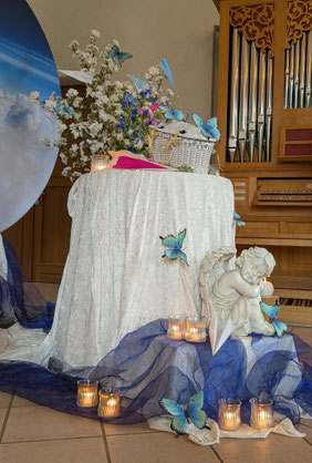 Mit weißen Decken geschmückten Altar, Frühsommerblumen in Blautönen, Schmetterlingen und Papierfliegern dekoriert. Auf dem Altar ein weißes Weidenkörbchen mit Deckel. Vor dem Altar sitzt eine Engelfigur umgeben von leuchtenden Teelichtgläsern