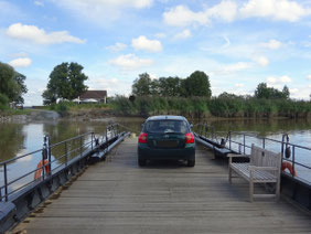 Ein dunkelgrüner Toyota steht auf dem hölzernen flachen Deck einer kleinen Fähre. Man sieht rechts und links die Reling, außerdem eine Holzbank. Am anderen Ufer die Zufahrt, hohes Schilf und ein Haus mit Sonnenschirmen davor.