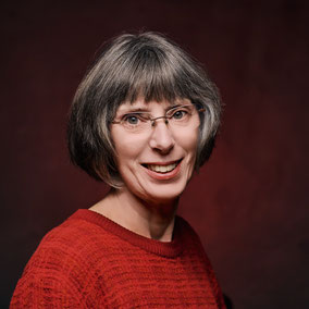 Een vrouw, Joke Zonneveld, voor een groene beukenhaag. Ze kijkt stralend uit haar ogen, draagt een bril, heeft een bruinkleurige sjaal om, en jas aan. Haar haar is bruin met grijstinten. 