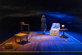 Photo du spectacle "Mamamé" : sur un plancher de bois, a droite une table basse et une boîte posée par terre, au fond à gauche un fauteuil et un guéridon, au centre de dos une comédienne en manteau vert et petit chapeau.