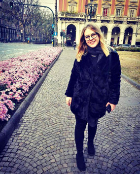 Foto von Aila Valjevac. Aila stht auf Kopfsteinpflaster in einer Stadt.Links sind rosa Blumen zu erkennen. Im Hintergrund befindet sich eine Ampel.