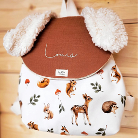 sac enfant avec deux oreilles de lapin en peluche le devant est en peluche couleur crème avec le rabat imprimé lapins dans des tons vert marron et gris le rabat est fini avec un passepoil doré