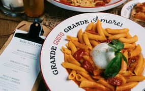 Ein Teller Rigatoni mit Tomatensauce, Burrata und Basilikum steht im Mittelpunkt, umgeben von Menükarte, weiteren Pastagerichten und Getränken auf einem rustikalen Restauranttisch.