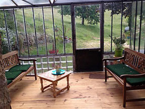 Intérieur de la véranda vitrée ouvrant sur la nature, une vallée avec des arbres, une table basse, un salon de rotin par gite de la gite de la gorre à louer en ardeche