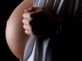 Körperauschnitt eines Babybauches  im Stil einer Mondsichel beleuchtet auf schwarzem Hintergrund, rechts hängt noch ein weißes Männerhemd, welches von einer männlichen Faust beiseite gezogen wird. Das Bild ist in schwarz weiß 