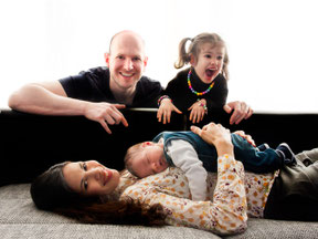 Dieses Foto zeigt ein Familienfoto. die Mutter liegt mit dem Baby auf dem Bauch auf einem Sofa. Der Vater und die Tochter schauen von hinter dem Sofa über die Lehne und haben Spaß.