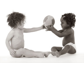 Ein hellhäutiges und ein dunkelhäutiges Baby reichen sich einen Ball auf einem weißen Studiohintergrund