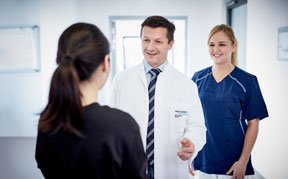 Ein Arzt und eine Krankenschwester begrüßen lächelnd eine Patientin in einem Krankenhausflur. 