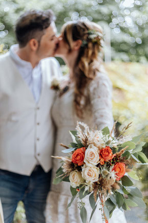 Der Fokus der Hochzeitsfotografin liegt hier auf dem rustikalen Brautstrauß. Das Brautpaar ist im Hintergrund sichtbar im Bokeh.