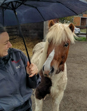 Sigrid und ihr Pferd Helgi stehen zusammen unter einem aufgespannten Regenschirm. Sigrid lacht, und nach Helgis Gesichtsausdruck zu schließen tut er das auch