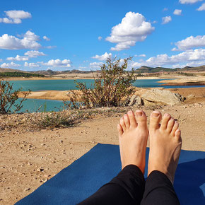 Auf dem Bild sind Füße auf einer Yogamatte zu sehen mit Blick auf einen See und blauen Himmel mit Wolken. Sina Emrich sitzt auf der Matte.