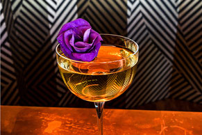 Milk Punch Godfather, der Cocktail ist in einem Nux Savage by Remi Savage Glas auf bernsteinfarbenem Tisch dekoriert mit einer wunderschönen blau-lila Blüte.