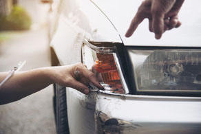 alt="Dunkles Auto mit frontalem Unfallschaden im Vordergrund, im Hintergrund begutachtet eine Person mit einem Laptop den Schaden."