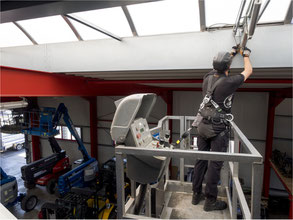 Person mit PSAgA (Helm, Auffanggurt und Höhensicherungsgerät) arbeitet gesichert aus einer Hubarbeitsbühne heraus an einem Hallendach