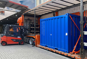 Man sieht einen komplett an der Seite geöffneten Planentieflader. Auf seiner Fläche steht eine containerartige Ware in Royalblau. Weiter hinten im Bild sieht man einen Stapler am Lkw.