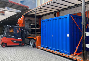 Man sieht einen komplett an der Seite geöffneten Planentieflader. Auf seiner Fläche steht eine containerartige Ware in Royalblau. Weiter hinten im Bild sieht man einen Stapler am Lkw.