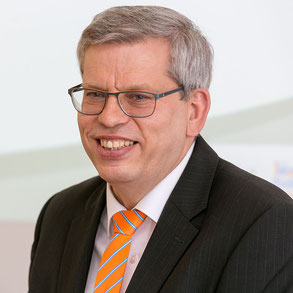 Roland Rüdinger, der Inhaber der Spedition, in leicht orangenem Hemd, mit blau/orangener Krawatte und schwarzem Jackett lächelt herzlich in die Kamera.