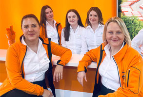 Eine Gruppenbild der Messe-Mannschaft auf der LogiMat. Man sieht Mitarbeiter und Mitarbeiterinnen in orangenem T-Shirt und Mitarbeiter und Mitarbeiterinnen in schwarzem Jackett und orangener Krawatte beziehungsweise Halstuch.