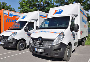 Zwei weiße Sprinter mit dem Firmenlogo stehen nebeneinander neben einem orangenen Lkw. Man blickt von der Vorderseite auf die beiden weißen Fahrzeuge der Spedition.