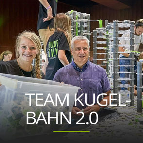 Lächelnde Frau hält eine transparente Kiste mit vielen Kunststoffteilen, Schriftzug: TEAM KUGELBAHN 2.0