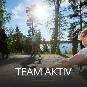 Menschen bei einer Teamübung neben einem See, Schriftzug: TEAM AKTIV