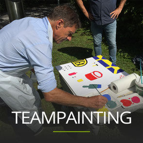 Mann bemalt einen Bilderrahmen in einer Wiese, Schriftzug: TEAMPAINTING