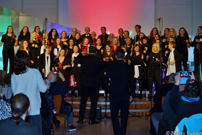 Blick aus dem Gang frontal auf die Bühne: Hanjo und Simon von hinten, UGC & Joyful Singers singen auf der Bühne