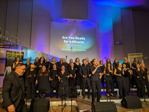 UGC singt schwarz gekleidet und in Bewegung, Hanjo dirigiert von links, Sophie singt vor dem Chor, im Hintergrund sind Liedtitel und blau-violette Lichter zu sehen