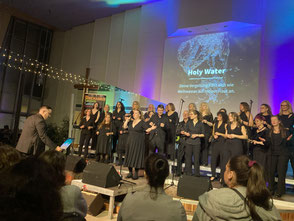 UGC singt schwarz gekleidet und groovend, Hanjo dirigiert auf der linken Bildseite, Rina singt vor dem Chor, im Hintergrund sind Liedtitel und blau-violette Lichter zu sehen