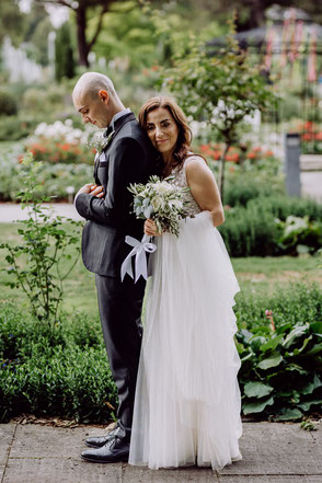 Braut im Brautkleid umarmt den Bräutigam von hinten, lehnt den Kopf an seine Schulterpartie und lächelt mit zur Seite geneigtem Kopf in die Kamera