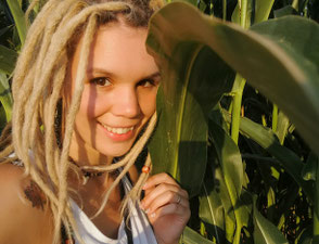 Meditationslehrerin Sabrina Sierks lächelt vor einem Maisfeld in die Kamera, ihre blonden Dreadlocks rahmen ihr Gesicht ein.