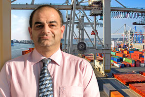 One of the Rüdinger employees smiles to the viewer, he fills the whole left half of the picture. He wears a pink shirt and a tie. On the right in the background you can see cranes in a port.