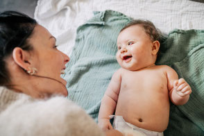 Mutter und Baby lachen gemeinsam, während das Baby auf einer Decke im Wohnzimmer liegt.