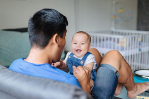 Baby sitzt auf dem Schoß des Vaters im Wohnzimmer und lacht fröhlich.