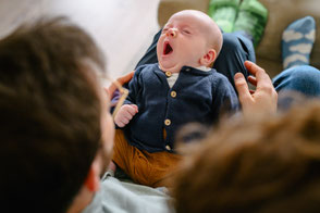 Baby liegt entspannt auf dem Schoß der Eltern im Wohnzimmer und gähnt.