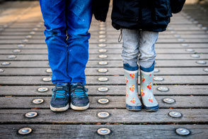 Geschwister stehen nebeneinander auf einer Holzbrücke, Fokus auf ihre Schuhe und Beine.