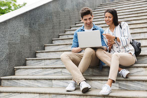Frau und Mann sitzen mit Laptop auf einer Treppe. Sie studieren im Ausland und sind mit einer Langzeit-Reiserücktritts-Versicherung der ERGO abgesichert.